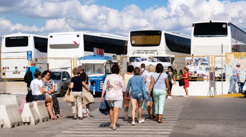Putnici peške prilaze turističkim autobusima na terminalu, prikaz organizovanog autobuskog prevoza i ukrcavanja za grčka letovališta
